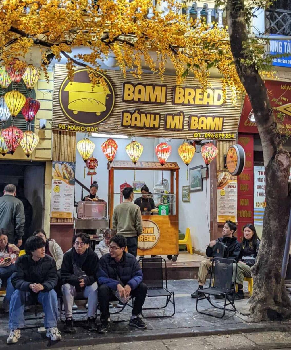 a crowd sitting on the sidewalk outside a banh mi vendor in hanoi