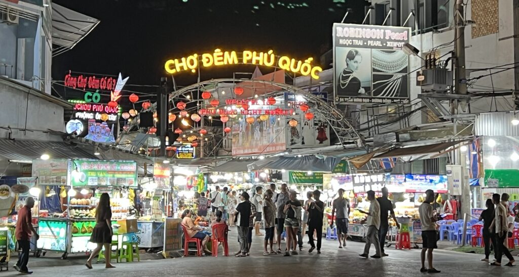 A bustling night market in Phú Quốc features brightly lit stalls and colorful signs under a large archway that reads "Chợ Đêm Phú Quốc." Crowds of people walk through the market, exploring various goods and food options. The vibrant atmosphere is highlighted by hanging red lanterns and illuminated shop displays.