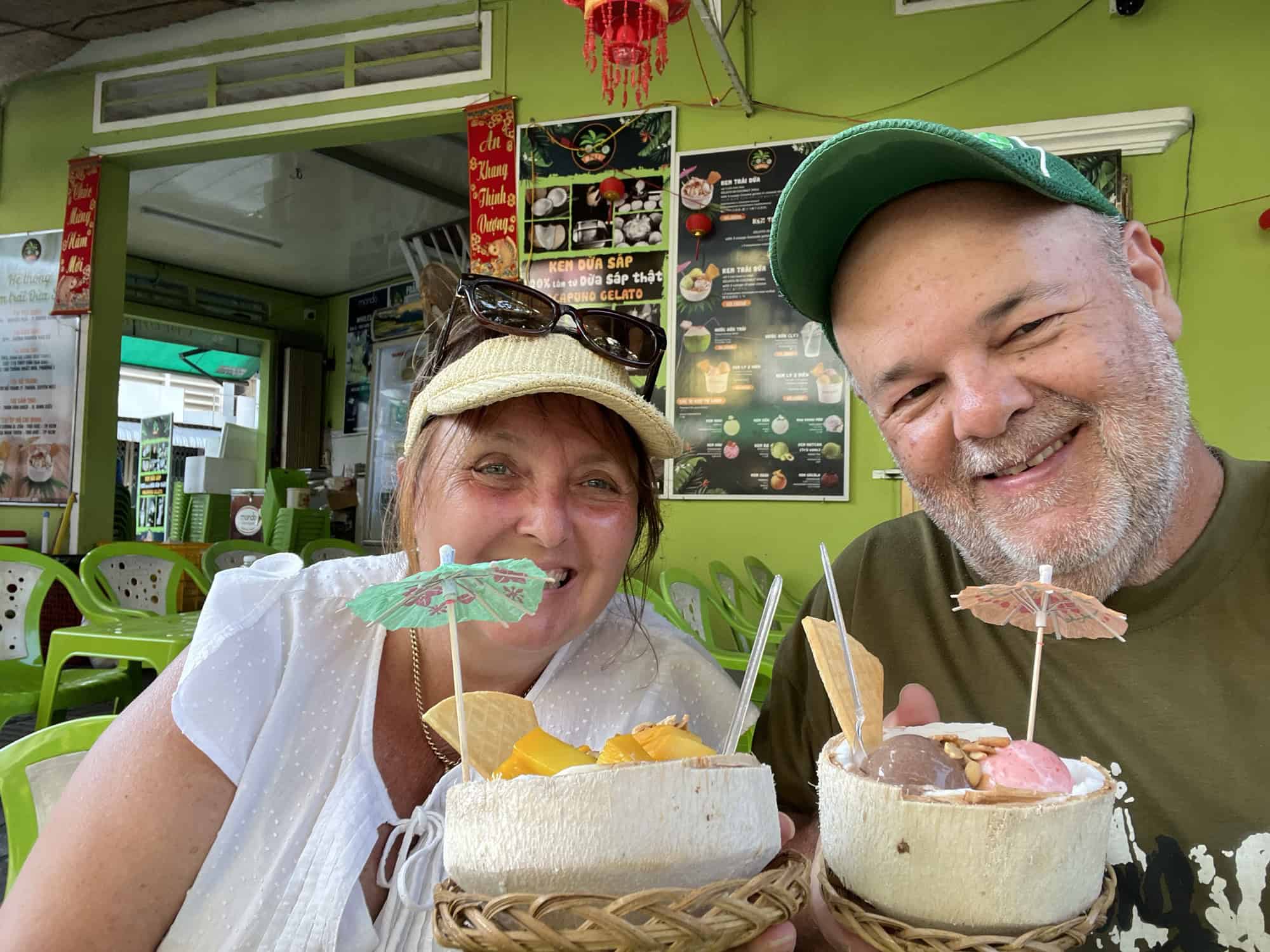 Dean and Pauline from Reasons to Visit sitting at an outdoor cafe in Phu Quoc, each holding a decorative coconut bowl filled with colorful ice cream and topped with paper umbrellas. The background shows green walls and a menu with various food options.