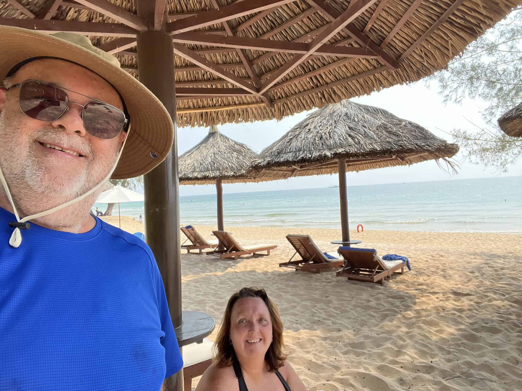 an and Pauline from Reasons to Visit at the beach in Phu Quoc, smiling under a thatched umbrella. Dean wears a wide-brimmed hat and sunglasses, while Pauline sits nearby with lounge chairs and more umbrellas visible behind them. The serene sea stretches out in the background.